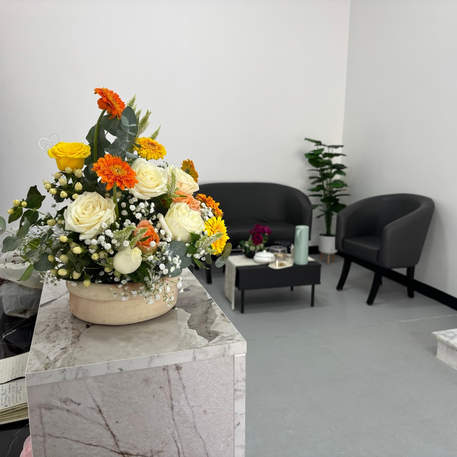 A flower arrangement on a marble surface, with grey waiting-room seating and a plant in the background